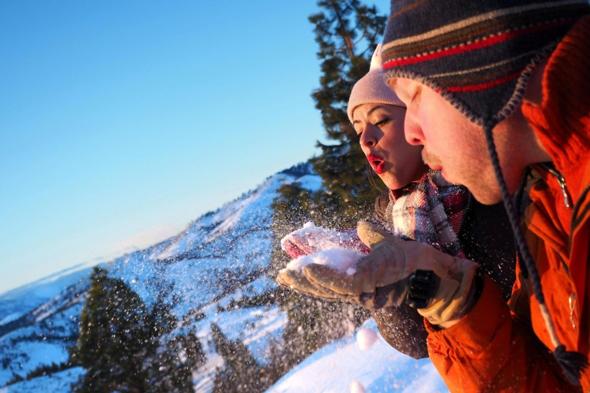 Gesichter zweier Personen in einer Winterlandschaft, die Schnee aus den Händen pusten