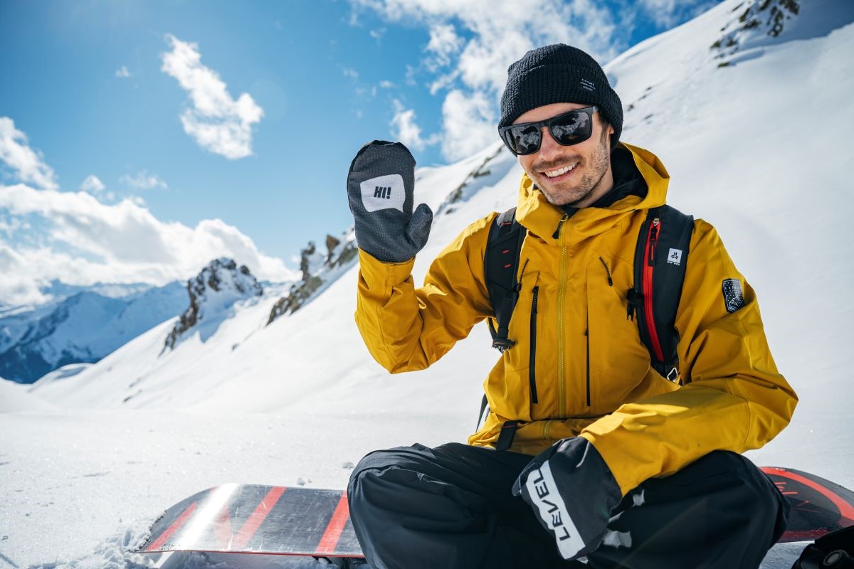 Thomas Feurstein sitzt auf einem Snowboard im Schnee und winkt mit der rechten Hand, im Hintergrund eine Piste und schneebedeckte Berge
