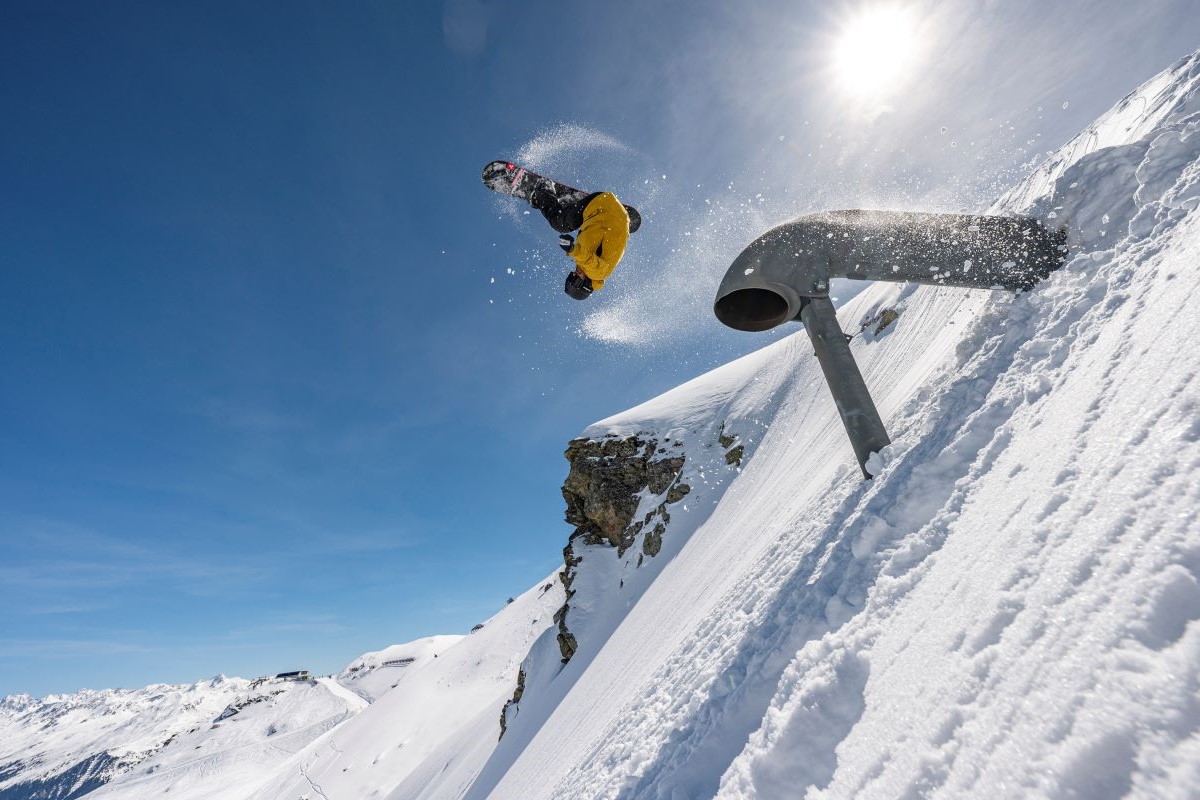 Thomas Feurstein fliegt mit seinem Snowboard kopfüber durch die Luft, der Hang ist schneebedeckt und im Hintergrund sind Berge zu sehen